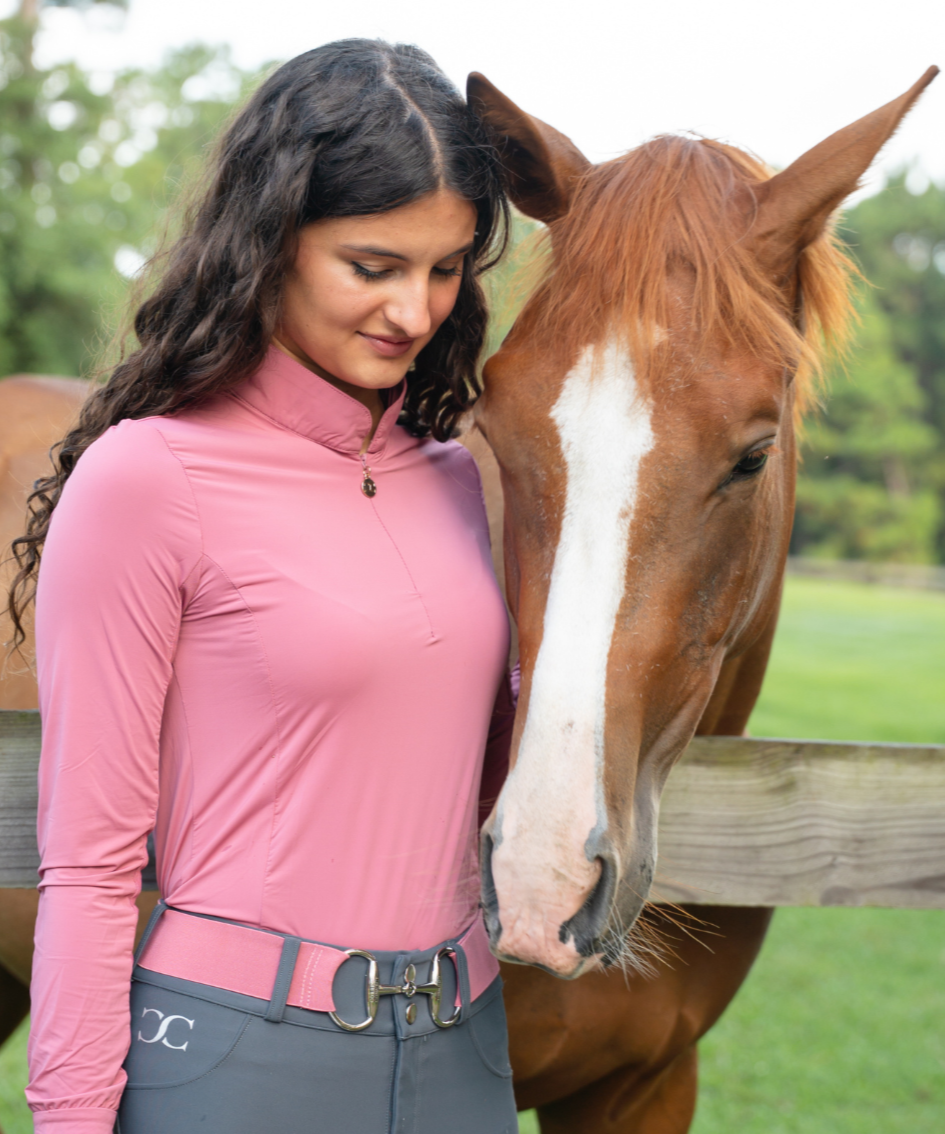 Woman in pink long-sleeve top and gray pants standing next to a brown horse with a white stripe on a wooden fence.